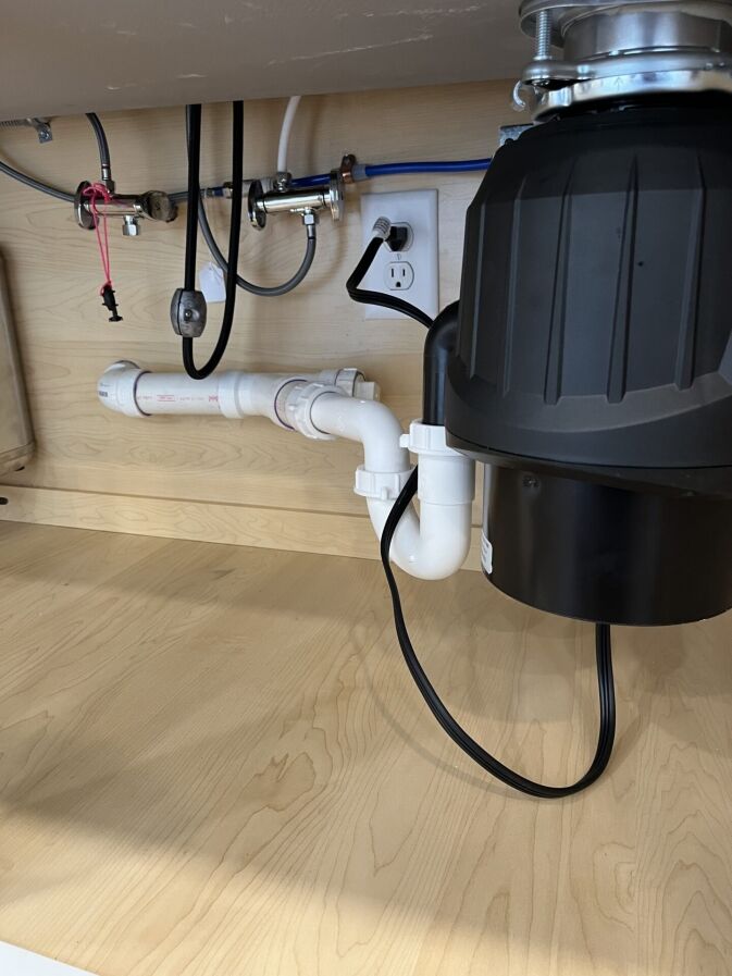 White PVC drain pipes and a black garbage disposal unit are installed under a sink; electrical outlet and cords are also visible against a wood cabinet backdrop.