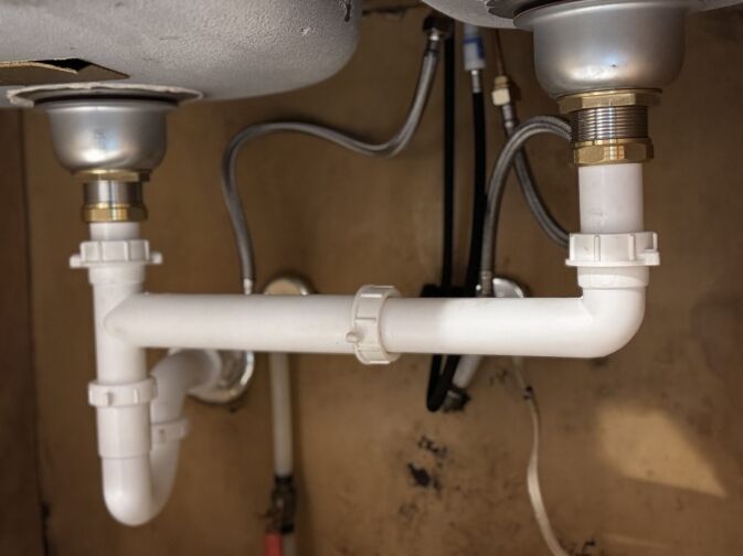 White PVC plumbing pipes and fittings are installed under a double sink, showing drain connections and water supply lines.