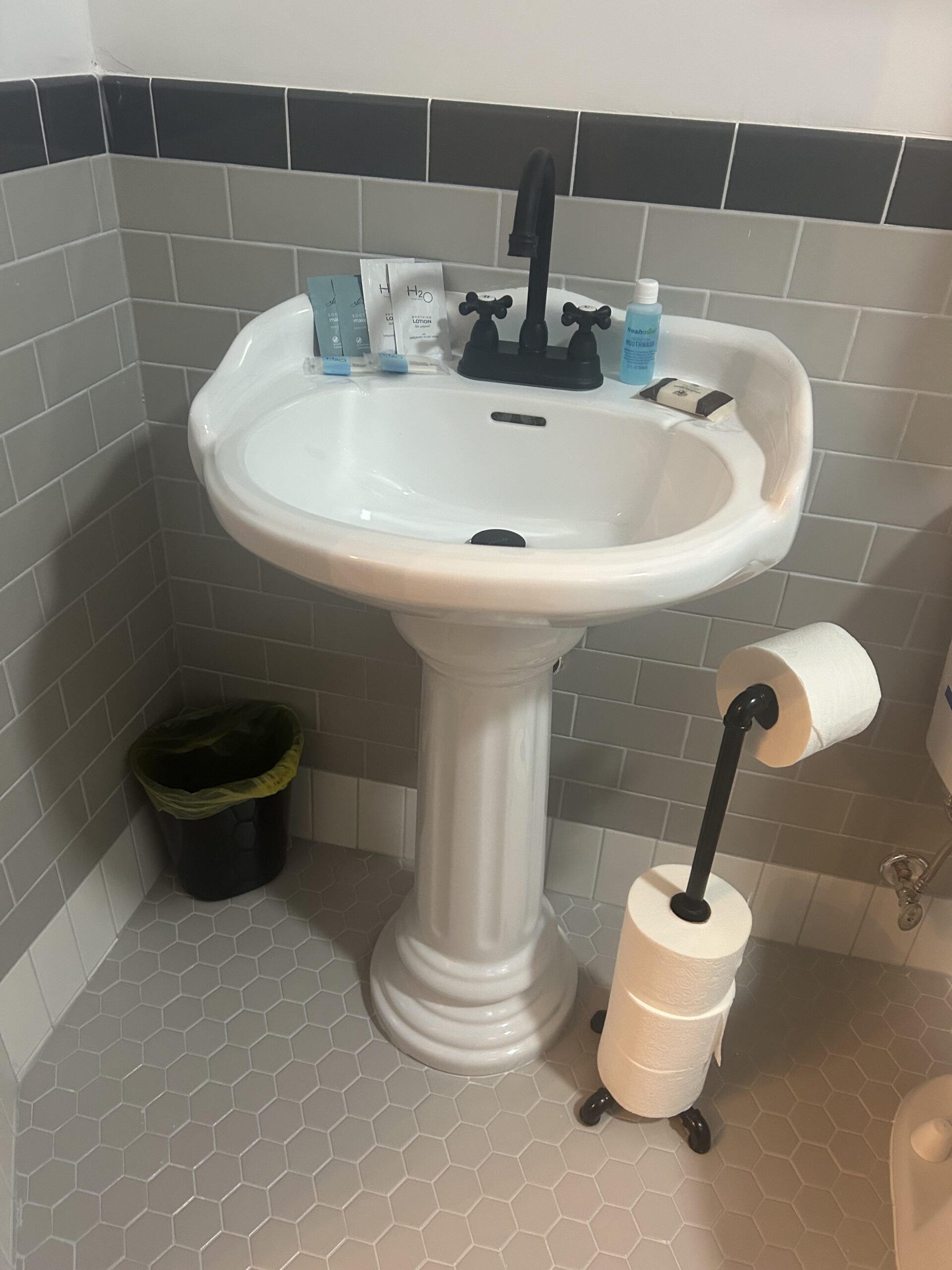 White pedestal sink with black faucet, toiletries on the back rim, a small trash can to the left, and a toilet paper stand with extra rolls on the right, in a tiled bathroom corner.