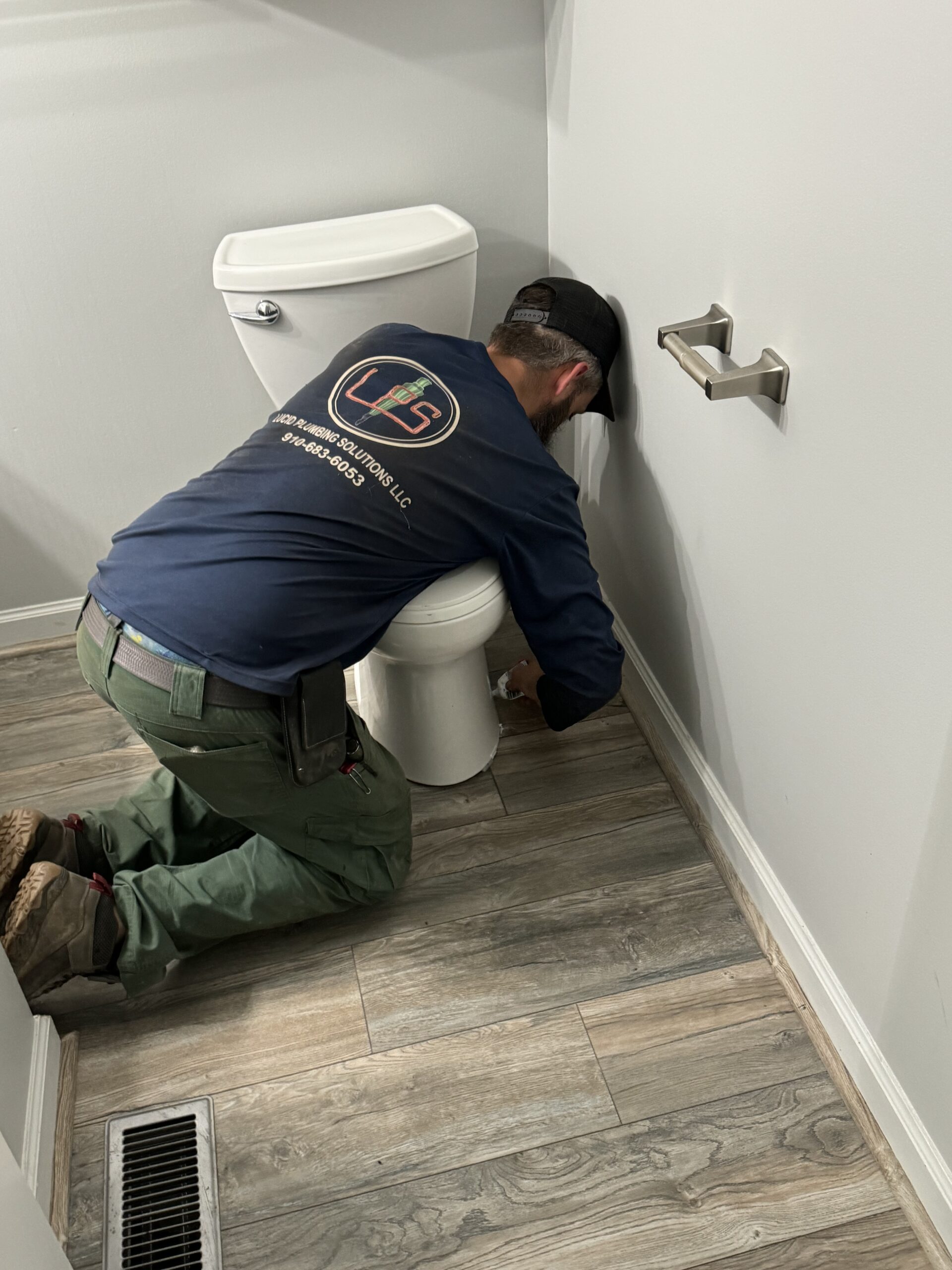 A plumber kneels on a bathroom floor beside a toilet, using tools to work near the toilet base.