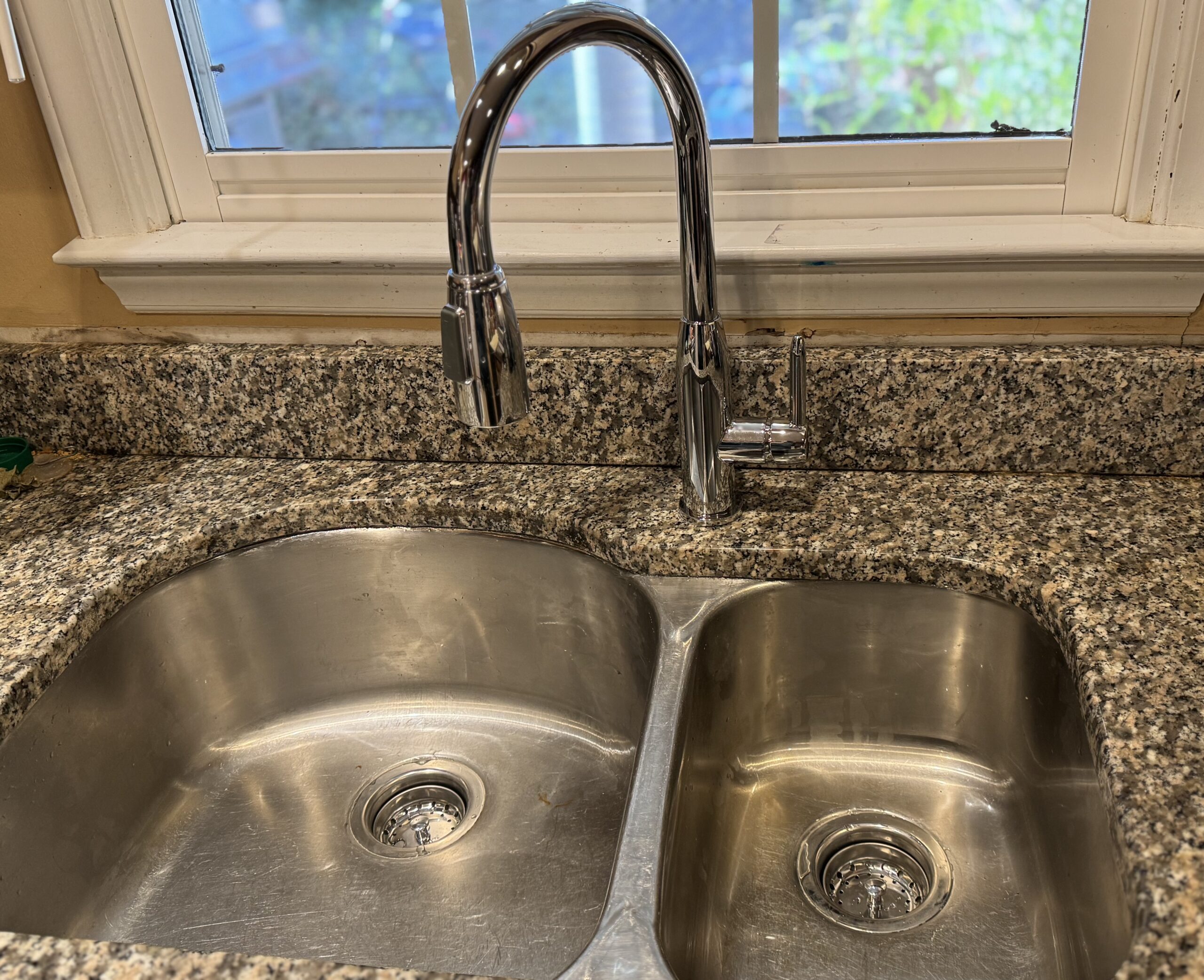 A double-basin stainless steel kitchen sink with a chrome faucet, set in a granite countertop below a window with white trim.