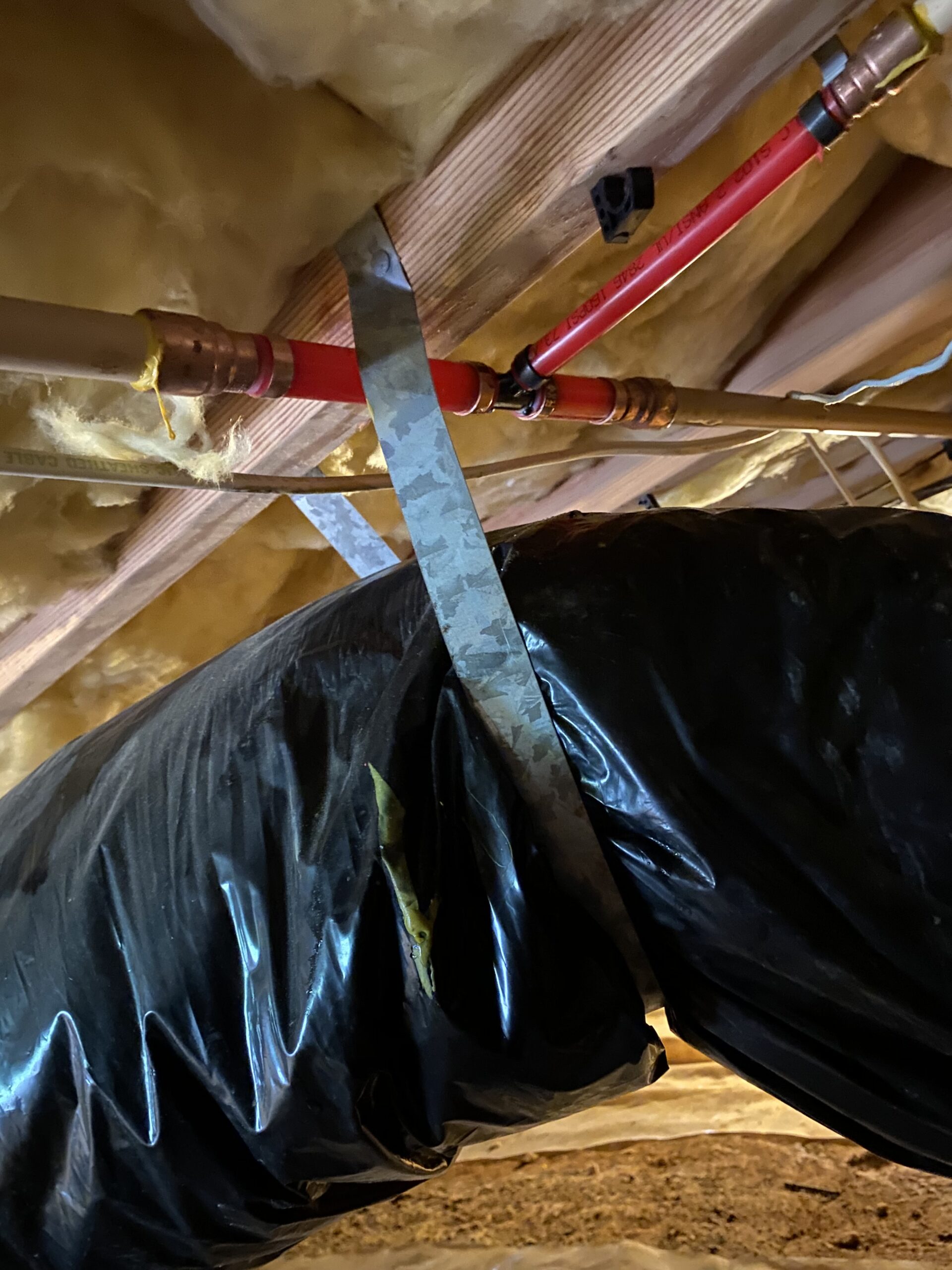 Exposed pipes and insulated duct secured with a metal strap under a wooden floor with yellow insulation above.