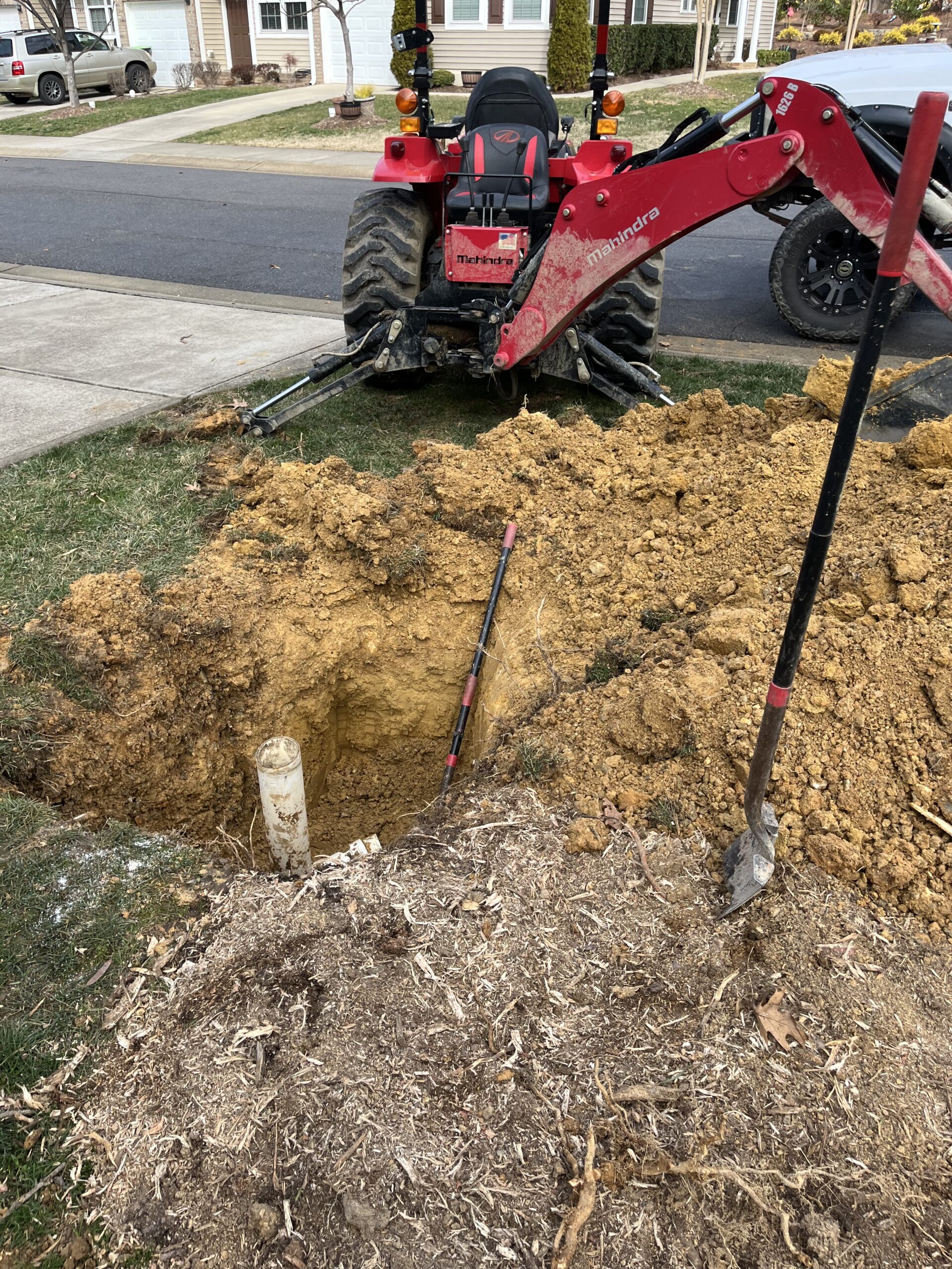 A backhoe tractor is parked next to a large, freshly dug hole in a yard near a sidewalk, with tools and piles of dirt and roots nearby.