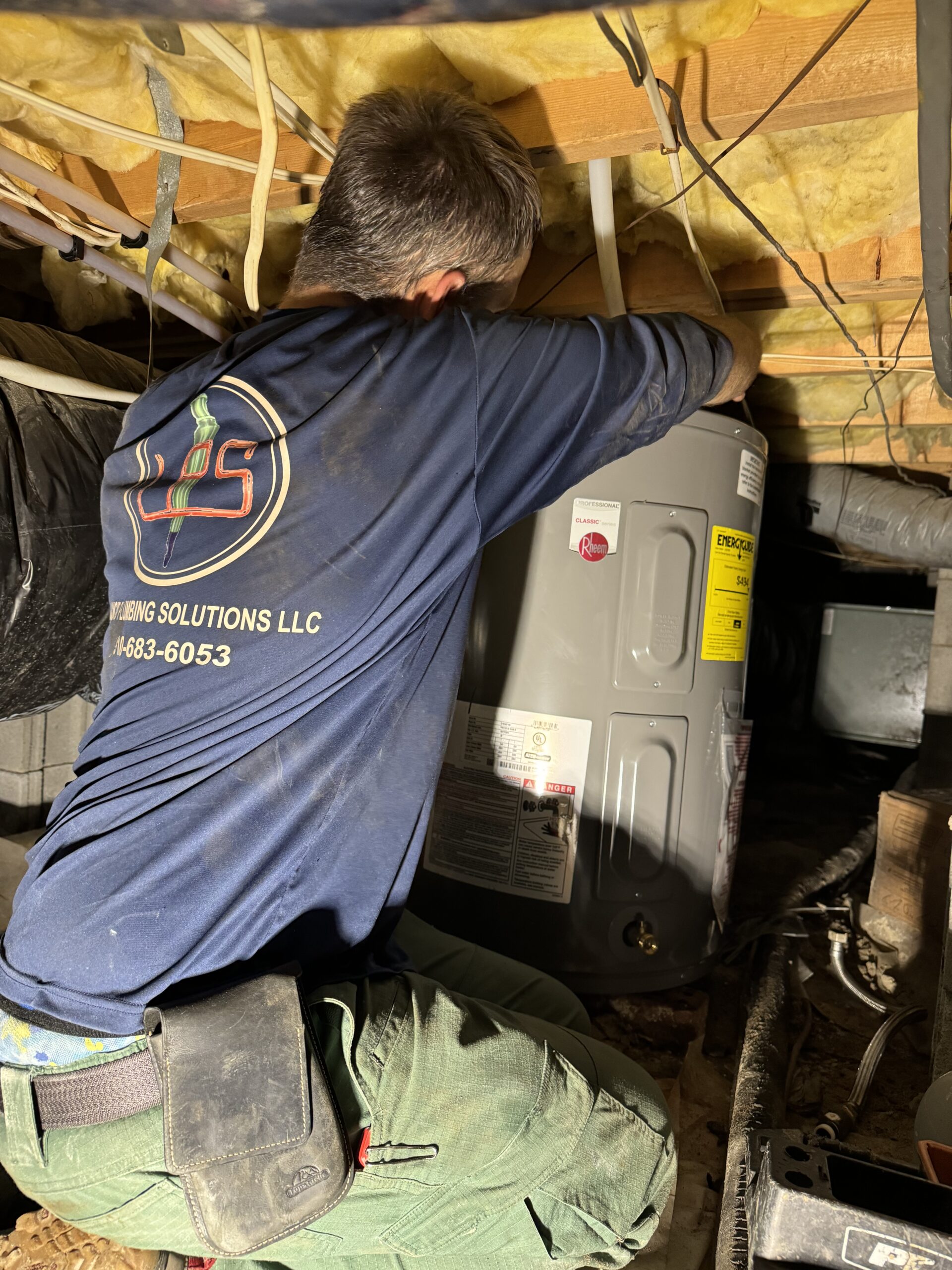A plumber in a blue shirt installs or repairs a water heater in a dimly lit crawl space with exposed insulation and pipes.