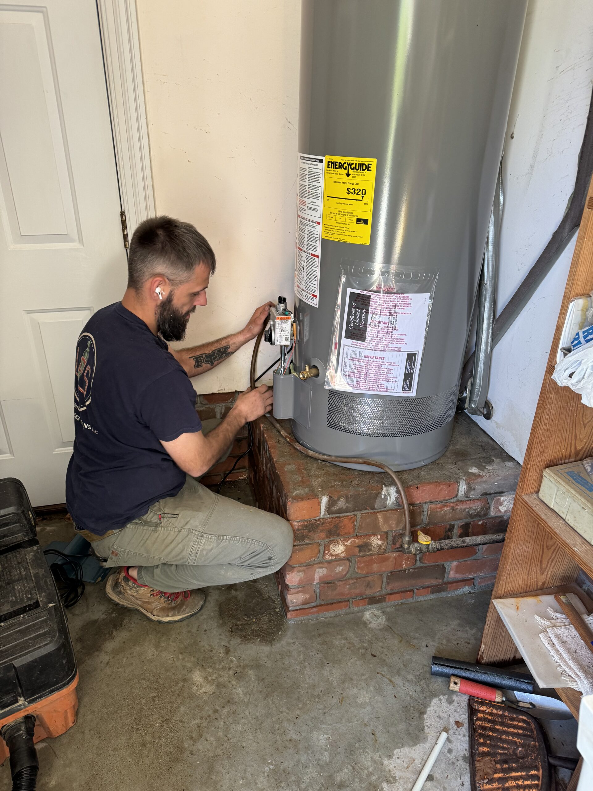A plumber kneels on the floor installing or repairing a water heater in a garage, using tools and wearing work clothes.