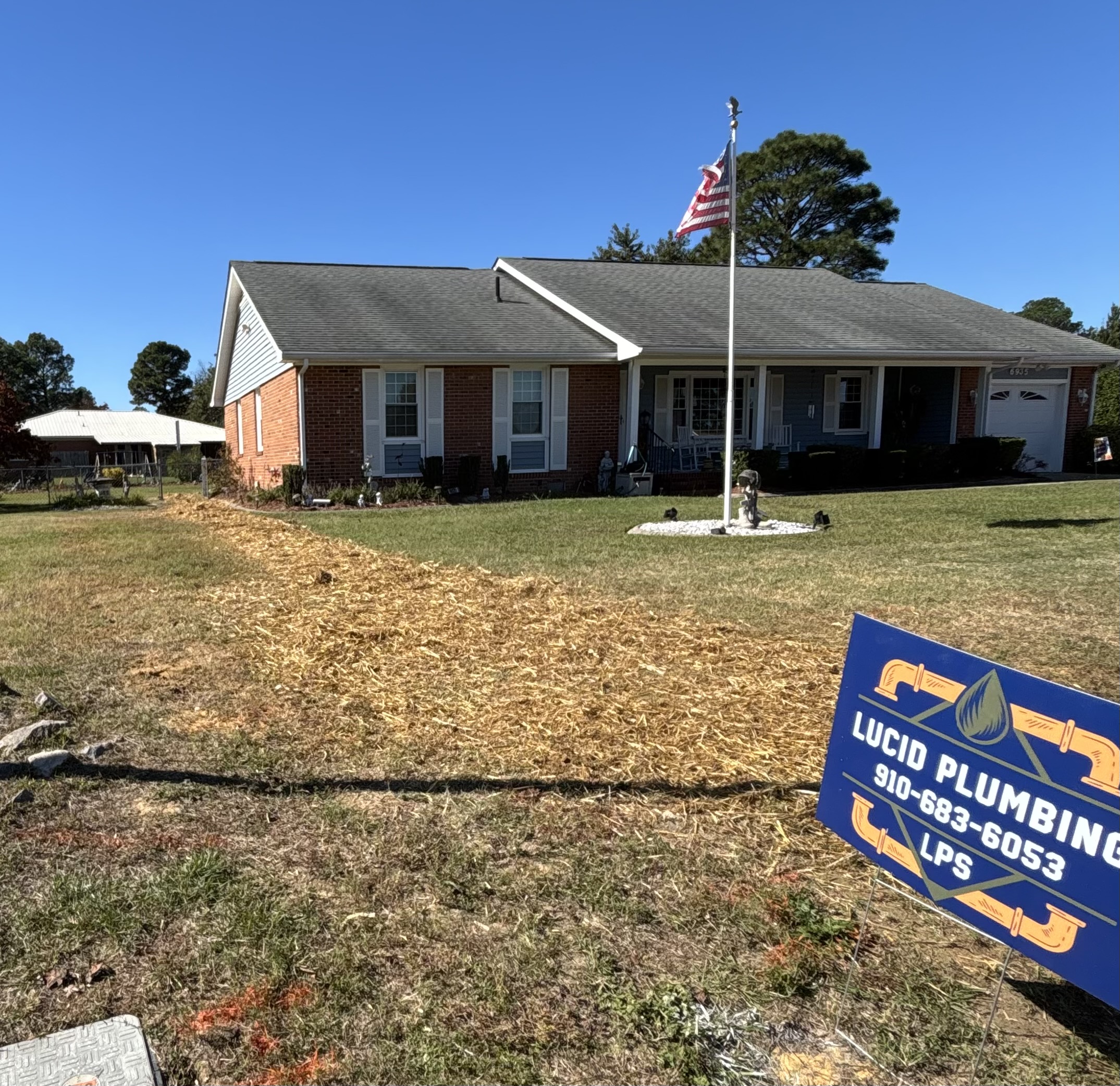 A single-story brick house with an American flag in the yard, a fresh trench filled with straw, and a Lucio Plumbing sign in the foreground.